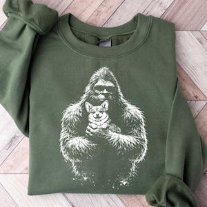 May include: Green sweatshirt featuring a white graphic of a gorilla holding a corgi. The sweatshirt has long sleeves and a crew neck. The graphic is centered on the front of the shirt. The fabric appears soft and comfortable.