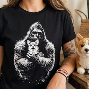 May include: Black t-shirt featuring a white graphic of a Bigfoot holding a Corgi dog. A plush Corgi toy sits on a wooden surface. The shirt has short sleeves and a crew neck.