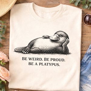Könnte beinhalten: Cremefarbenes T-Shirt mit einer Schwarz-Weiß-Illustration eines Schnabeltiers, das sich ausruht und den Kopf auf seine Hand stützt. Der Text "BE WEIRD. BE PROUD. BE A PLATYPUS." ist unter dem Schnabeltier-Bild gedruckt.