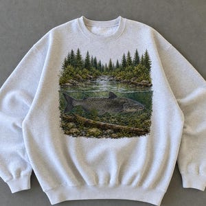 Può includere: Una felpa girocollo grigio chiaro con una stampa grafica dettagliata di un pesce che nuota sott'acqua in un fiume. La scena raffigura una foresta di pini verdi sopra la linea dell'acqua e un letto del fiume roccioso sotto la superficie.
