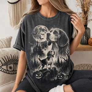 May include: A charcoal grey short-sleeve t-shirt featuring a graphic print of a beagle dog, a large full moon, flying bats, a dark castle silhouette, a forest, and three jack-o'-lanterns. The design is rendered in a monochromatic black and white style.