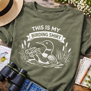 Puede incluir: Camiseta de manga larga verde oliva con el texto "THIS IS MY BIRDING SHIRT". El gr&aacute;fico muestra un p&aacute;jaro con prism&aacute;ticos en el agua con juncos. Tambi&eacute;n se ven un sombrero de paja, unos prism&aacute;ticos y un cuaderno.