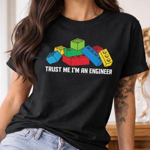 May include: Black t-shirt featuring a colorful graphic of toy building blocks in red, green, blue, and yellow. The text "TRUST ME I'M AN ENGINEER" is printed in white below the blocks.
