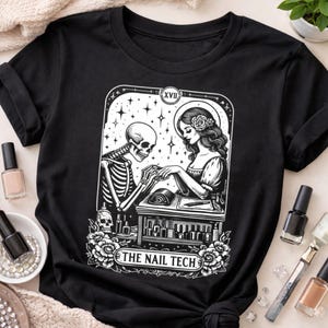 May include: Black t-shirt featuring a black and white graphic of a skeleton giving a manicure to a woman. The design includes the text "THE NAIL TECH" and is framed by floral and star elements. Nail polish bottles are visible.