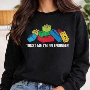 Peut inclure: Sweat-shirt noir avec un motif coloré de blocs de construction rouges, verts, bleus et jaunes. Le texte "TRUST ME I'M AN ENGINEER" est imprimé en dessous des blocs en blanc.