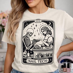 Könnte beinhalten: Cremefarbenes T-Shirt mit einer Schwarz-Weiß-Grafik, die ein Skelett zeigt, das einer Frau eine Maniküre macht. Das Design umfasst Sterne, einen Mond und den Text "NAIL TECH". Das Bild hat eine Tarotkarten-Ästhetik.