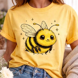 May include: Yellow t-shirt featuring a cartoon bee with large eyes, wings, and black and yellow stripes. The bee is surrounded by small star shapes. The shirt has short sleeves and a rolled hem.