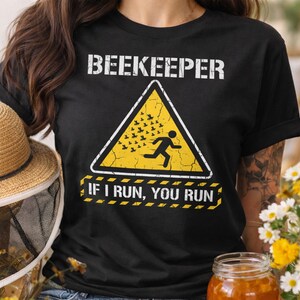 以下が含まれることがあります： 「BEEKEEPER」の文字が書かれた黒いTシャツ。黄色い危険標識の上に、走る人物と蜂が描かれています。「IF I RUN, YOU RUN」のテキストが下にあります。麦わら帽子と花が背景にあります。