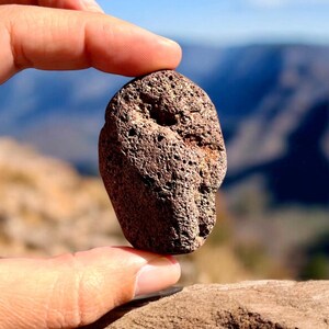 Könnte beinhalten: Nahaufnahme eines dunkelbraunen, rauen und strukturierten Steins mit einem Loch in der Mitte. Der Stein wird von einer Hand vor einem verschwommenen Hintergrund aus Bergen und Himmel gehalten.