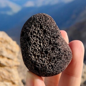 May include: A dark gray, rough, porous rock with a textured surface. The rock is being held in a hand against a blurry background of mountains.