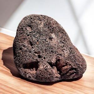 Może przedstawiać: Szary, chropowaty, porowaty kamień o zaokrąglonym kształcie. Kamień leży na jasnobrązowej drewnianej powierzchni.