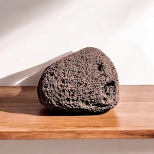 May include: A dark brown, porous lava rock with an irregular shape. The rock sits on a wooden surface against a white background. The rock's texture is rough and full of small holes.