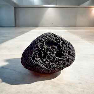 Könnte beinhalten: Ein großer, poröser Stein in dunkelgrau mit einer rauen, unebenen Oberfläche. Der Stein liegt auf einem hellgrauen Boden.