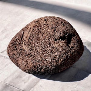 Könnte beinhalten: Ein poröser, dunkelbrauner Lavastein mit rauer, strukturierter Oberfläche. Der Stein hat eine unregelmäßige, abgerundete Form und ist wahrscheinlich ein dekoratives oder natürliches Element.
