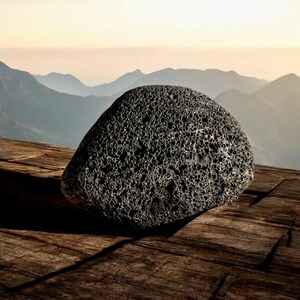 May include: A dark gray, porous rock with a rough texture sits on a wooden surface. The rock is in focus, while the background is blurred and shows a mountain range in the distance.