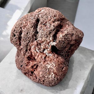 May include: A reddish-brown lava rock with a porous texture and an irregular shape. The surface is covered in small holes and a few white mineral deposits. The rock's appearance suggests a natural, geological formation.