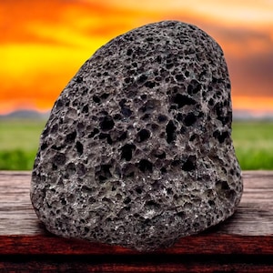 Könnte beinhalten: Ein dunkelgrauer, poröser Bimsstein mit zahlreichen Löchern. Der Stein ist vor einem verschwommenen Hintergrund eines Sonnenuntergangs über einem grünen Feld platziert. Der Stein liegt auf einer Holzoberfläche.