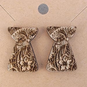 May include: Two brown, intricately carved earrings with a floral design are displayed on a tan card. The earrings have a unique shape, wider at the bottom and narrower at the top. The background includes a rainbow-coloured fabric.