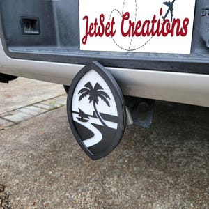 May include: A black and white trailer hitch cover depicting a tropical island scene with a palm tree, boat, and water. The cover has a black border and is attached to a vehicle. The text "JetSet Creations" is visible.