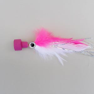 May include: A pink and white fishing lure with a silver eye and pink feathers. The lure has a cylindrical pink head and a cluster of white and pink feathers with silver tinsel.