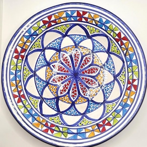 May include: A large, round ceramic plate with a colourful geometric design. The plate features a central motif of a flower with blue, red, yellow, and green petals. The border of the plate is decorated with a repeating pattern of triangles and squares in various colours.