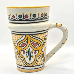 May include: A white ceramic mug with a yellow, red and blue geometric pattern. The mug has a handle and is decorated with a repeating pattern of dots. The text "STYLE 4" is visible below the mug.