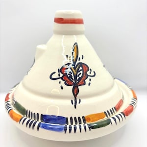 May include: A cream-colored ceramic tagine pot with a conical lid and a circular base. The lid features a red band and a central floral design in red, yellow, and blue. The base is decorated with colorful stripes.