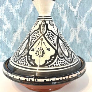 May include: A black and white ceramic tagine pot with a conical lid and a round base. The tagine features intricate black patterns on a white background. The base is a reddish-brown colour. The text "Big Tajine Black & White" is at the bottom.