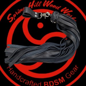 May include: Black leather flogger with a braided handle and a silver-colored clasp. The image features the text "Spring Hill Wood Works Handcrafted BDSM Gear" in red on a black background.