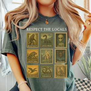 May include: Moss green t-shirt featuring a graphic of cryptids like Bigfoot, Loch Ness Monster, and Alien, with the text "RESPECT THE LOCALS". The design is printed on a comfortable, casual tee.