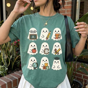 May include: A light green t-shirt featuring a repeating pattern of cartoon ghosts with school-themed accessories. Each ghost holds items like an apple, books, a pencil, and a globe. The shirt is paired with a white pleated skirt.