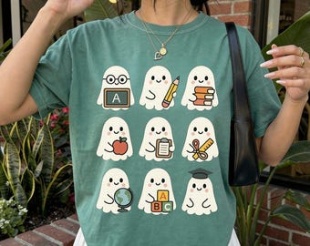 May include: A light green t-shirt featuring a repeating pattern of cartoon ghosts with school-themed accessories. Each ghost holds items like an apple, books, a pencil, and a globe. The shirt is paired with a white pleated skirt.
