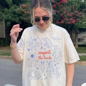 May include: Ivory-colored t-shirt featuring blue illustrations of mythical creatures and an alien, with the text "respect the locals" in red. The shirt is a relaxed fit.