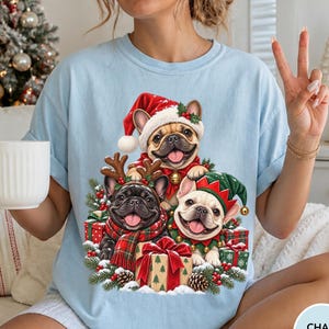 May include: A light blue t-shirt featuring a graphic of three French bulldogs wearing Christmas attire, including a Santa hat, reindeer antlers, and an elf hat, surrounded by wrapped gifts, pine branches, and holly berries.