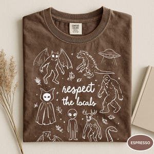 May include: Espresso-colored t-shirt with a crew neck. The shirt features a white graphic of various cryptids and a UFO, with the text "respect the locals". The shirt is made of a soft material.