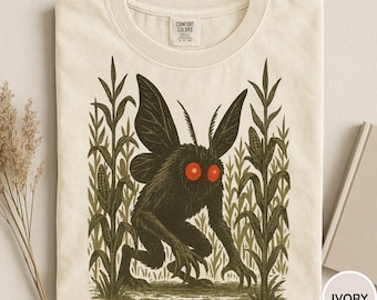 May include: Ivory-colored t-shirt featuring a dark green cryptid creature with red eyes, large wings, and sharp claws, standing in a cornfield. The shirt has a Comfort Colors tag.