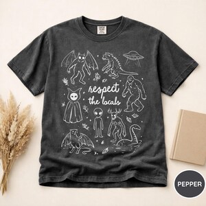 May include: Dark gray t-shirt with a white graphic featuring various cryptids and the text "respect the locals." The design includes a bat-like creature, Godzilla, a UFO, Bigfoot, an alien, and the Loch Ness Monster.