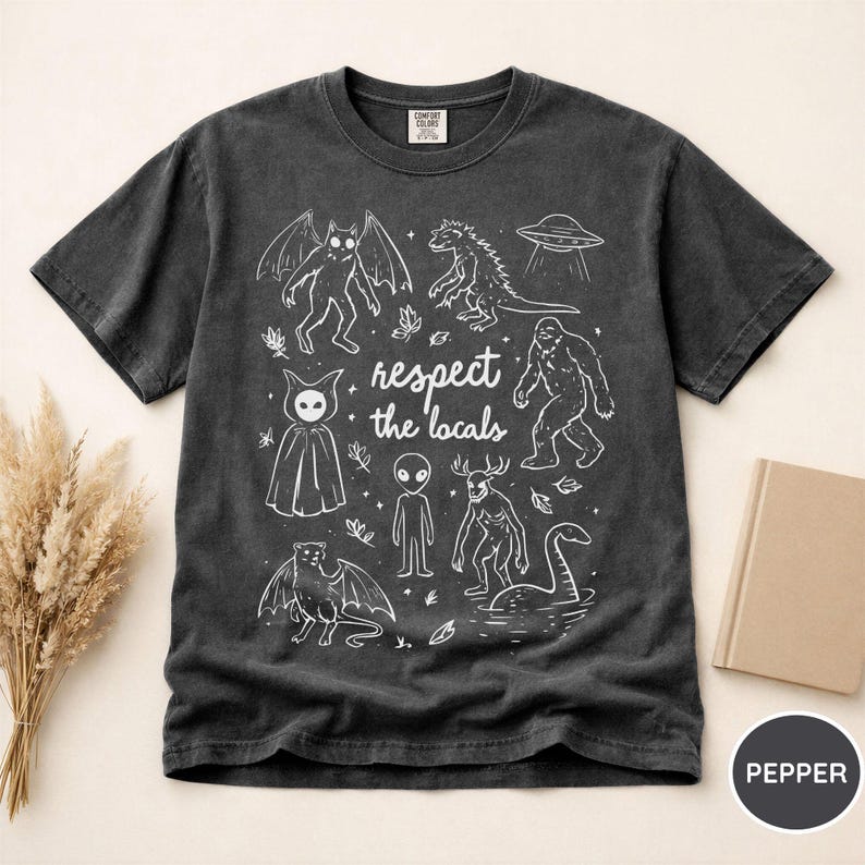 May include: Dark gray t-shirt with a white graphic featuring various cryptids and the text "respect the locals." The design includes a bat-like creature, Godzilla, a UFO, Bigfoot, an alien, and the Loch Ness Monster.