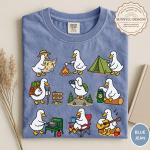 May include: A blue crew neck t-shirt featuring a graphic print of nine white ducks engaged in various camping activities, including reading a map, roasting a marshmallow, setting up a tent, hiking, using binoculars, holding a lantern, cooking, sitting in a chair, and playing guitar.