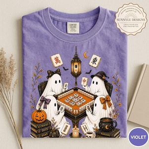 May include: A violet t-shirt featuring a graphic of four ghosts playing mahjong at a table. The design includes a hanging lantern, a pumpkin, stacks of books, and mahjong tiles. The shirt has a Comfort Colors label at the inner neckline.