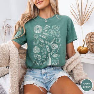 Puede incluir: Una camiseta de manga corta de color verde claro con un estampado gr&aacute;fico de l&iacute;neas blancas de varias formas de pasta, incluyendo farfalle, ravioli y espaguetis, junto con una botella y un tenedor, dispuestos en un patr&oacute;n disperso.