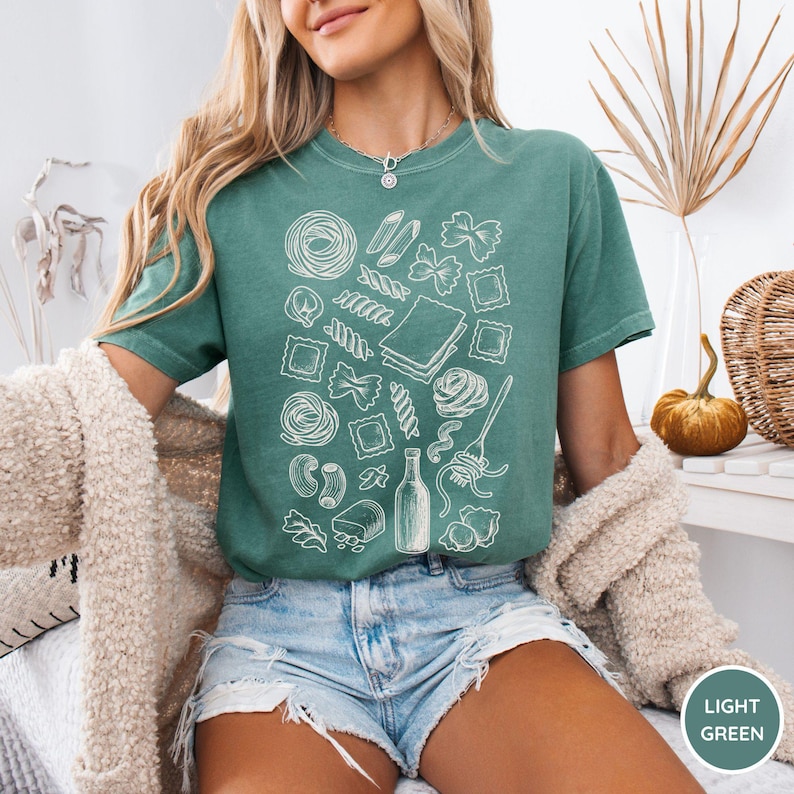 Puede incluir: Una camiseta de manga corta de color verde claro con un estampado gr&aacute;fico de l&iacute;neas blancas de varias formas de pasta, incluyendo farfalle, ravioli y espaguetis, junto con una botella y un tenedor, dispuestos en un patr&oacute;n disperso.