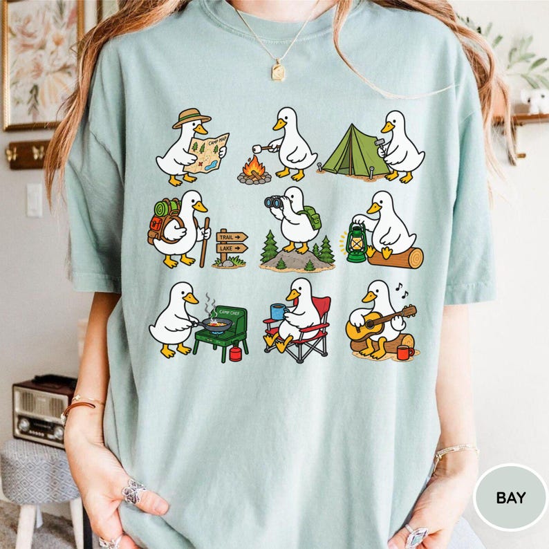 May include: A light sage green t-shirt featuring a graphic print of nine white ducks engaged in various camping activities, including hiking with a map, roasting marshmallows, setting up a tent, cooking on a stove, and playing a guitar.
