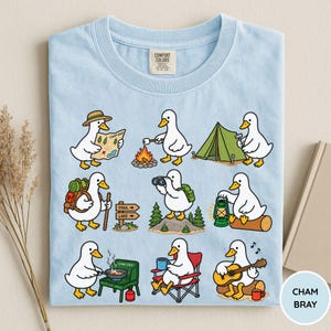 May include: A light blue t-shirt featuring a graphic print of white ducks engaged in various camping activities, including reading a map, roasting marshmallows, setting up a tent, hiking, using binoculars, holding a lantern, cooking, and playing guitar.