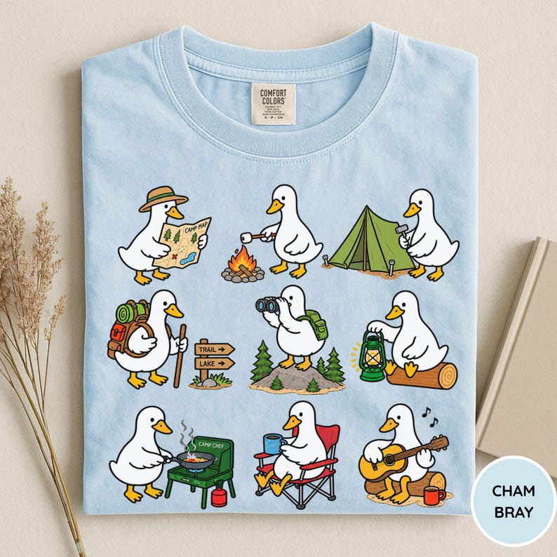 May include: A light blue t-shirt featuring a graphic print of white ducks engaged in various camping activities, including reading a map, roasting marshmallows, setting up a tent, hiking, using binoculars, holding a lantern, cooking, and playing guitar.