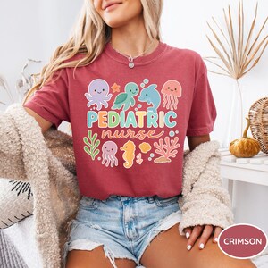 K&ouml;nnte beinhalten: Karmesinrotes T-Shirt mit dem Schriftzug "Pediatric Nurse" in bunten Buchstaben, umgeben von Cartoon-Meeresbewohnern. Das Design umfasst einen Oktopus, eine Schildkr&ouml;te, einen Delfin, eine Qualle, ein Seepferdchen und Korallen.