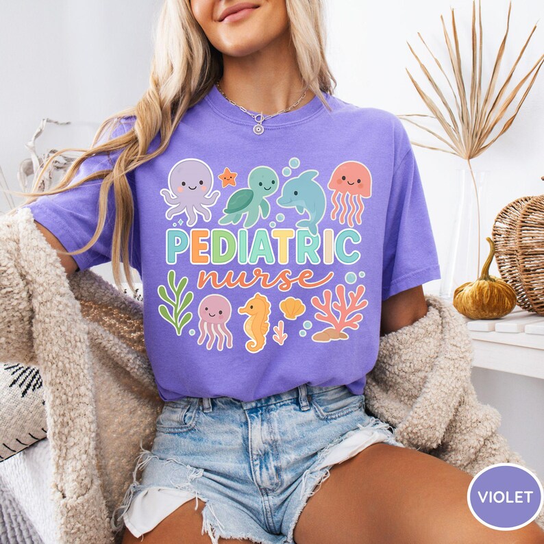 K&ouml;nnte beinhalten: Ein violettes T-Shirt mit dem Schriftzug "PEDIATRIC nurse" und einem farbenfrohen Design mit Meeresbewohnern. Das Design umfasst einen Oktopus, eine Schildkr&ouml;te, einen Delfin, eine Qualle, ein Seepferdchen und Korallen. Das Shirt ist aus weichem Material.