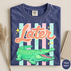 May include: A navy blue t-shirt with a graphic design. The design features the word "Later" in orange script, a green alligator, and vertical stripes in pink and teal. Yellow stars are also included.