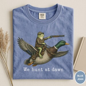 Puede incluir: Camiseta azul vaquero con una ilustraci&oacute;n humor&iacute;stica de una rana con ropa de cazador montada en un pato y sosteniendo una escopeta. El texto "We hunt at dawn" est&aacute; impreso debajo. La camiseta est&aacute; doblada.