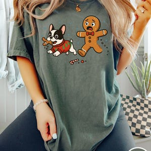 May include: A moss green t-shirt featuring a cartoon illustration of a French bulldog wearing a Christmas sweater, holding a bone, and a scared gingerbread man. The shirt has a distressed look and a round neckline.